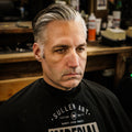 Before shot of a man seated in a barber chair wearing an Imperial barber cape, with silver hair slicked back but unstyled, in a barbershop setting.