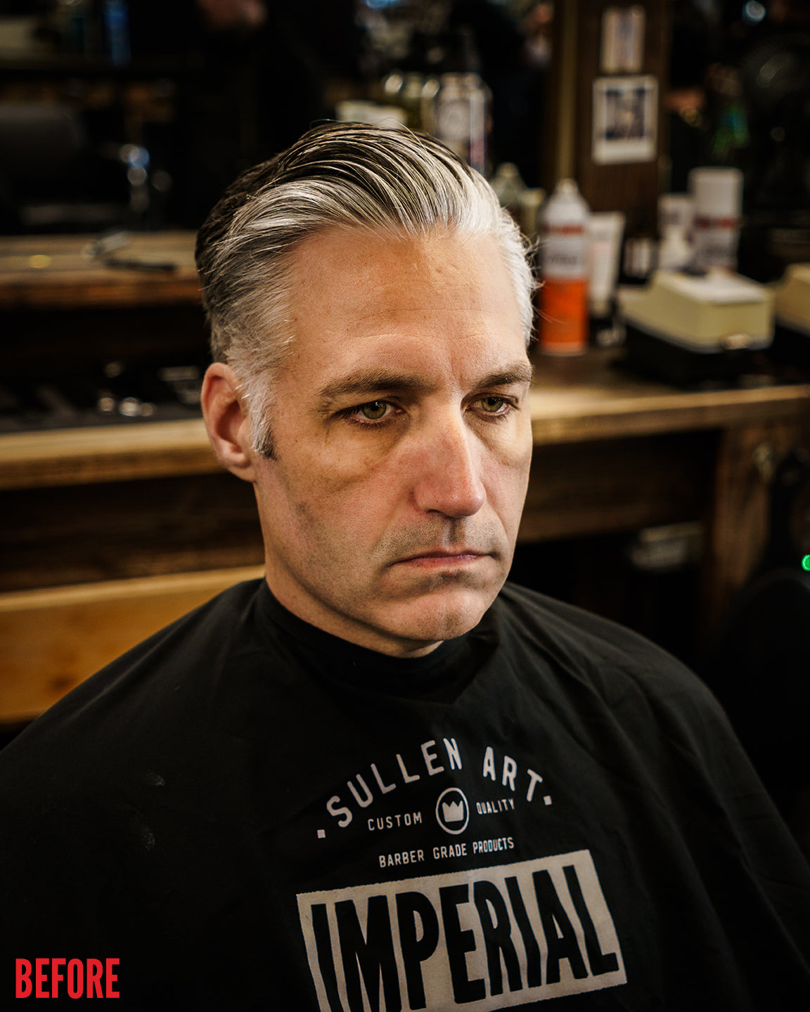 Before shot of a man seated in a barber chair wearing an Imperial barber cape, with silver hair slicked back but unstyled, in a barbershop setting.