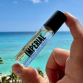 A hand holds the Imperial Barber Grade Products Crown Roll cologne bottle against a bright turquoise ocean backdrop.