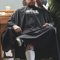 A tattooed man seated in a barber chair wearing the black Imperial Barber Grade Products cape while a barber works on his fade.